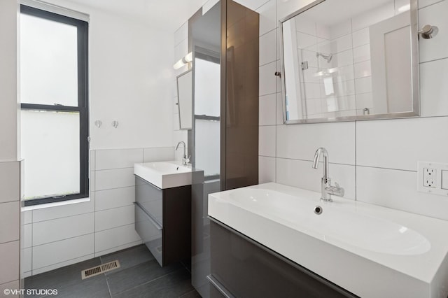 full bath featuring a tile shower, tile walls, two vanities, and visible vents