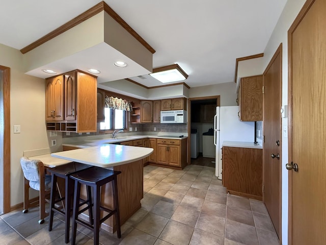 kitchen with a peninsula, a sink, tasteful backsplash, white appliances, and brown cabinetry