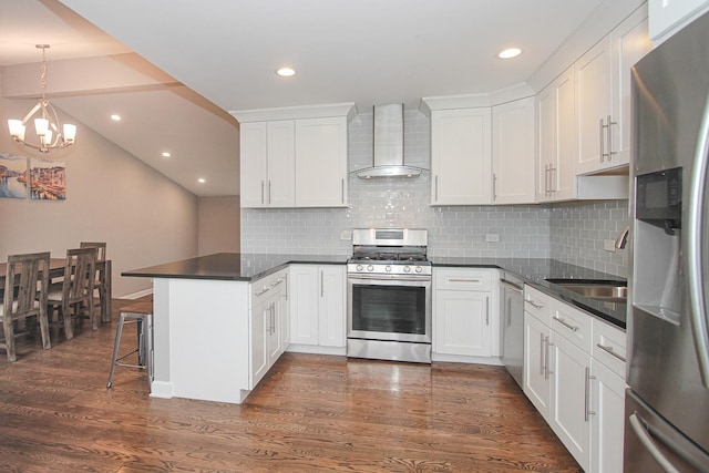 kitchen featuring stainless steel appliances, wall chimney exhaust hood, a peninsula, a notable chandelier, and dark wood-type flooring