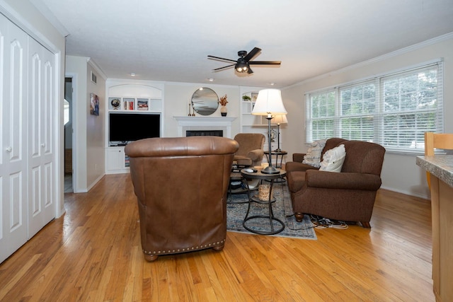 living area featuring ornamental molding, built in shelves, a ceiling fan, a tiled fireplace, and light wood-style floors