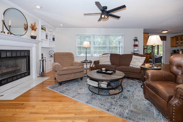 living room with ornamental molding, built in shelves, light wood-style flooring, a ceiling fan, and a tiled fireplace