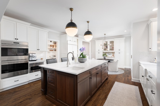 kitchen with white cabinets, dark brown cabinets, stainless steel double oven, a sink, and a kitchen island with sink