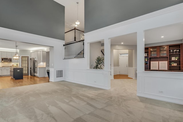 unfurnished living room featuring light carpet, visible vents, a towering ceiling, stairs, and a decorative wall