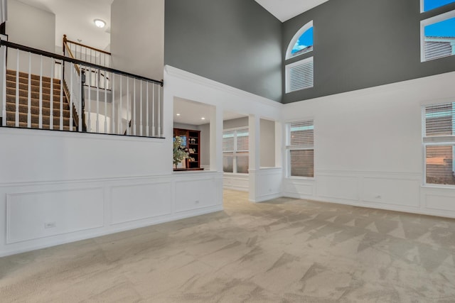 unfurnished living room featuring a towering ceiling, a wainscoted wall, carpet floors, a decorative wall, and stairs