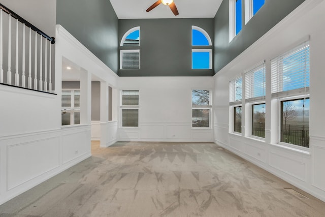 unfurnished living room featuring a towering ceiling, a wainscoted wall, a decorative wall, carpet flooring, and ceiling fan