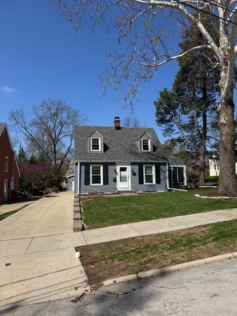 cape cod home with a front lawn, driveway, a shingled roof, and a chimney