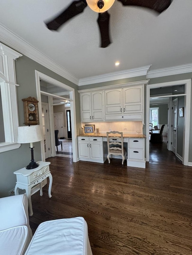 living area featuring a ceiling fan, built in study area, baseboards, dark wood-style floors, and ornamental molding