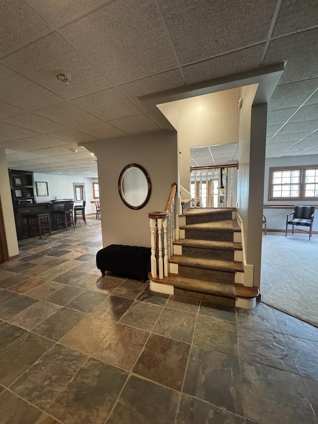 staircase with bar area, a drop ceiling, and stone tile floors