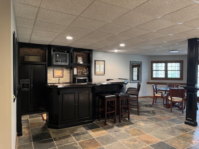 bar featuring black refrigerator with ice dispenser, stone tile floors, wet bar, baseboards, and wall oven