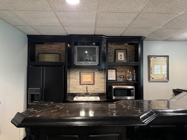 bar featuring black fridge with ice dispenser, stainless steel microwave, a drop ceiling, and indoor bar