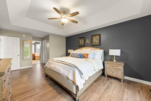bedroom featuring a raised ceiling, baseboards, wood finished floors, a ceiling fan, and ensuite bathroom