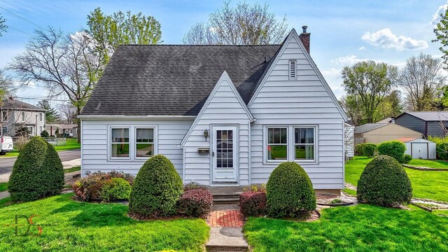 view of front of home with a front lawn, a chimney, a shingled roof, a storage shed, and an outbuilding