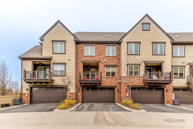 multi unit property featuring brick siding, driveway, cooling unit, a garage, and roof with shingles