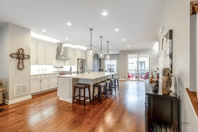 kitchen with an island with sink, a kitchen breakfast bar, wood finished floors, wall chimney range hood, and stainless steel fridge