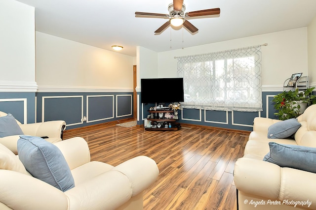 living area featuring a decorative wall, wainscoting, wood finished floors, and a ceiling fan