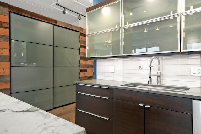 kitchen featuring a sink, dark brown cabinetry, backsplash, glass insert cabinets, and light stone countertops