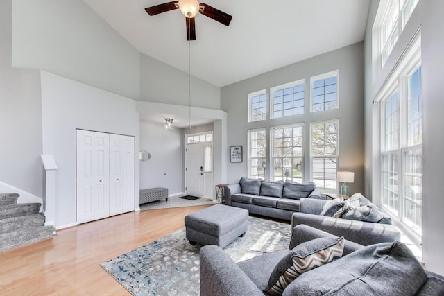 living area featuring stairs, light wood-style floors, baseboards, high vaulted ceiling, and a ceiling fan