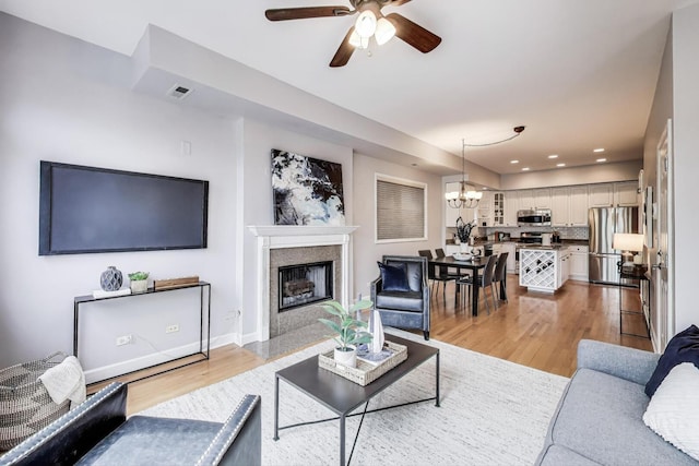living area featuring light wood-style floors, a tiled fireplace, ceiling fan with notable chandelier, baseboards, and visible vents