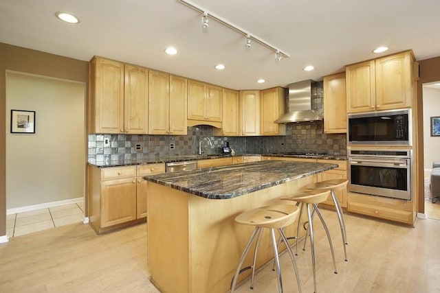 kitchen with light brown cabinetry, wall chimney range hood, baseboards, appliances with stainless steel finishes, and a breakfast bar area