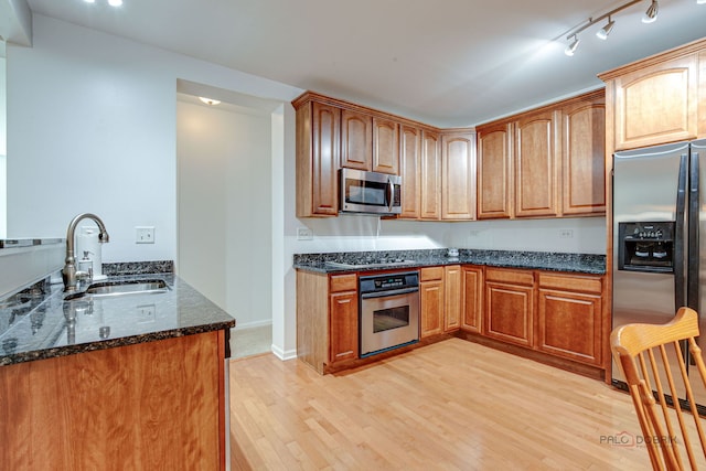 kitchen featuring appliances with stainless steel finishes, brown cabinets, a sink, light wood finished floors, and dark stone countertops