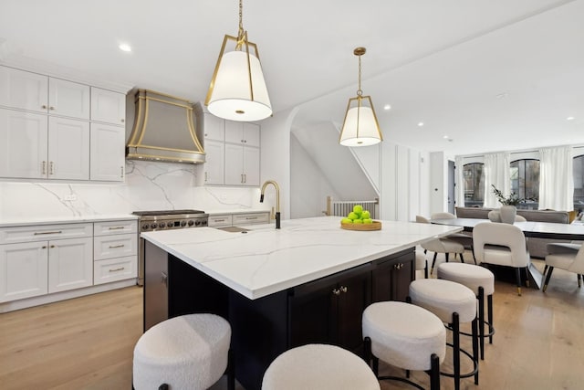 kitchen featuring premium range hood, light wood-style flooring, a sink, a center island with sink, and backsplash