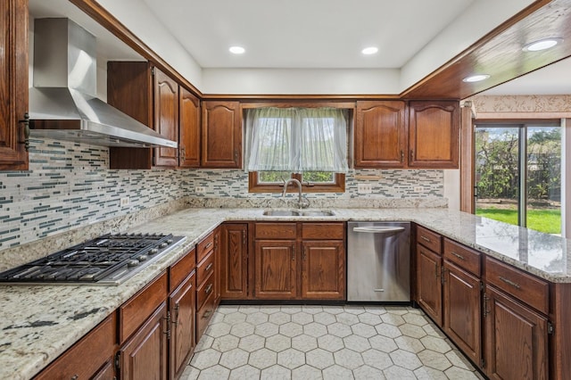 kitchen with a peninsula, wall chimney range hood, light stone counters, a sink, and appliances with stainless steel finishes