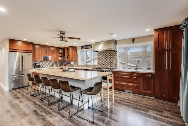 kitchen with high quality appliances, wall chimney range hood, dark wood-style floors, a kitchen breakfast bar, and light countertops