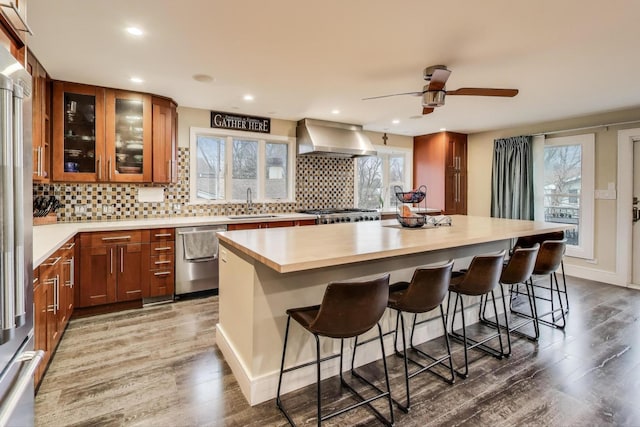 kitchen with appliances with stainless steel finishes, wall chimney range hood, light countertops, brown cabinetry, and wood finished floors