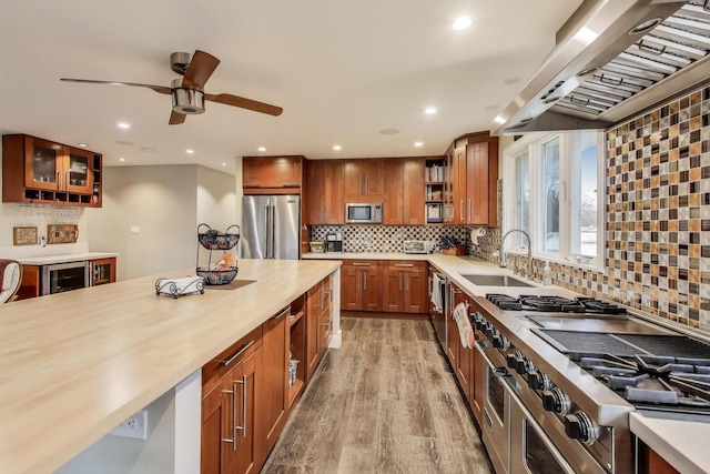 kitchen with high quality appliances, a sink, light wood finished floors, glass insert cabinets, and wall chimney range hood