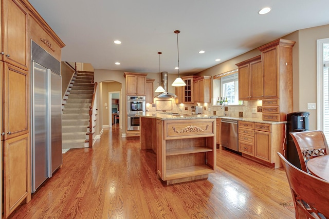 kitchen featuring wall chimney range hood, decorative backsplash, appliances with stainless steel finishes, arched walkways, and open shelves
