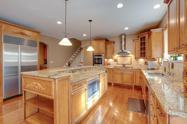 kitchen featuring wall chimney range hood, a kitchen island, a sink, light wood-style floors, and built in appliances