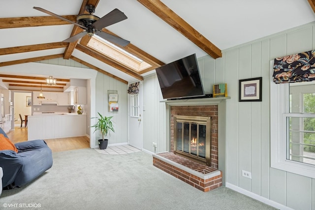 living area featuring ceiling fan, light colored carpet, a brick fireplace, baseboards, and lofted ceiling with skylight