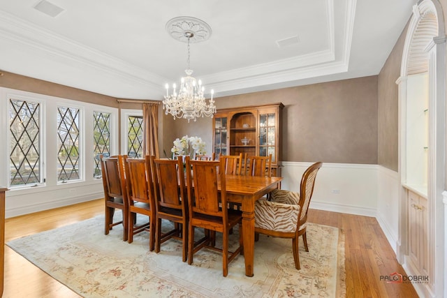 dining room featuring light wood-style flooring, a tray ceiling, wainscoting, a chandelier, and crown molding