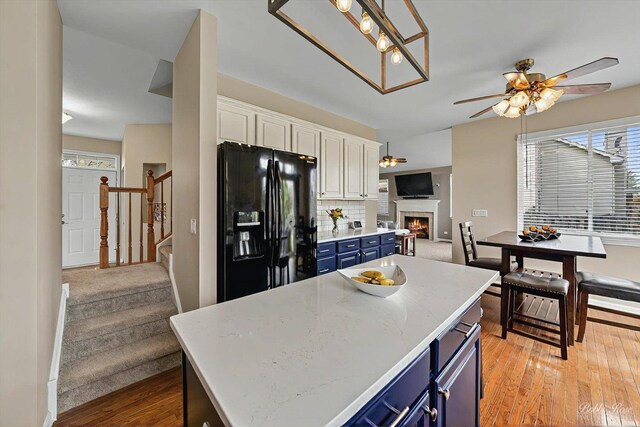 kitchen featuring black fridge with ice dispenser, a ceiling fan, a warm lit fireplace, and blue cabinetry