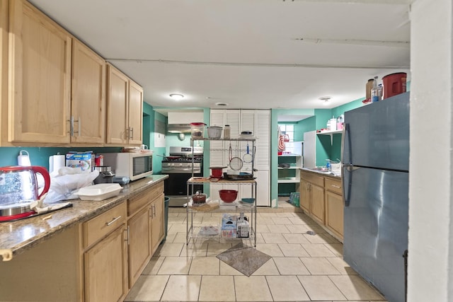 kitchen featuring light brown cabinetry, light tile patterned flooring, freestanding refrigerator, gas stove, and white microwave