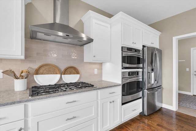 kitchen featuring wall chimney range hood, backsplash, appliances with stainless steel finishes, dark wood-style floors, and white cabinetry
