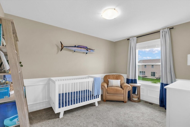 bedroom featuring visible vents, wainscoting, a crib, carpet, and a decorative wall
