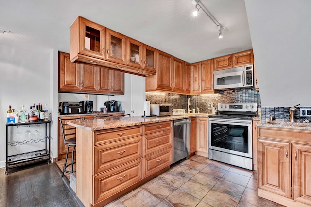 kitchen with light stone counters, glass insert cabinets, appliances with stainless steel finishes, rail lighting, and backsplash