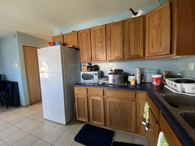kitchen with a sink, light tile patterned flooring, brown cabinetry, dark countertops, and freestanding refrigerator