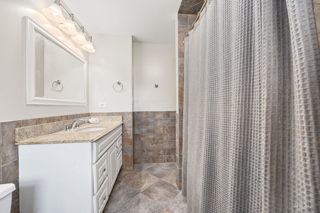 full bath featuring stone finish floor, a wainscoted wall, a shower with shower curtain, tile walls, and vanity