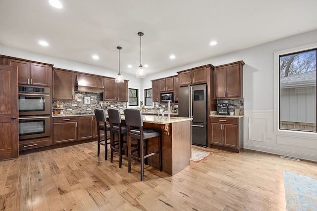 kitchen featuring a kitchen breakfast bar, light wood-style flooring, dark brown cabinetry, appliances with stainless steel finishes, and premium range hood