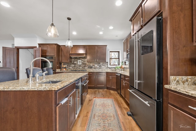 kitchen with a sink, tasteful backsplash, light wood-type flooring, a center island with sink, and appliances with stainless steel finishes