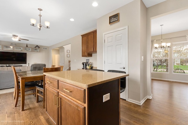 kitchen with wood finished floors, decorative light fixtures, a kitchen island, light stone countertops, and ceiling fan with notable chandelier