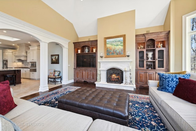 living area with light wood-type flooring, plenty of natural light, vaulted ceiling, decorative columns, and a lit fireplace