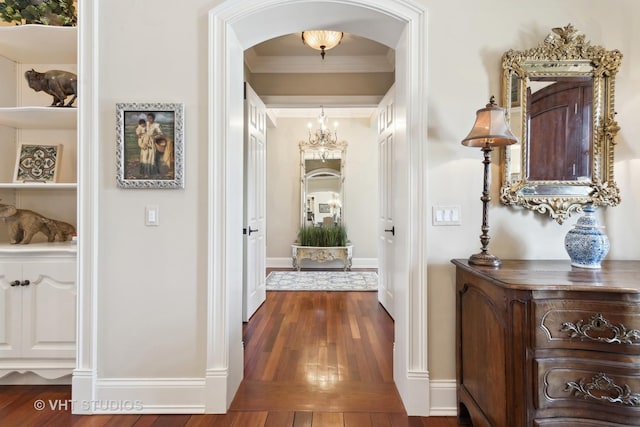 corridor with dark wood-style floors, arched walkways, baseboards, a notable chandelier, and crown molding