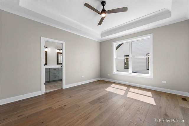 unfurnished bedroom with wood finished floors, baseboards, ensuite bath, ornamental molding, and a raised ceiling