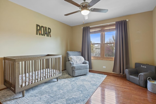 bedroom with ceiling fan, a nursery area, hardwood / wood-style flooring, and baseboards