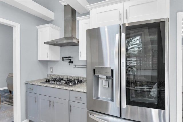 kitchen with appliances with stainless steel finishes, wall chimney range hood, white cabinets, light stone countertops, and baseboards