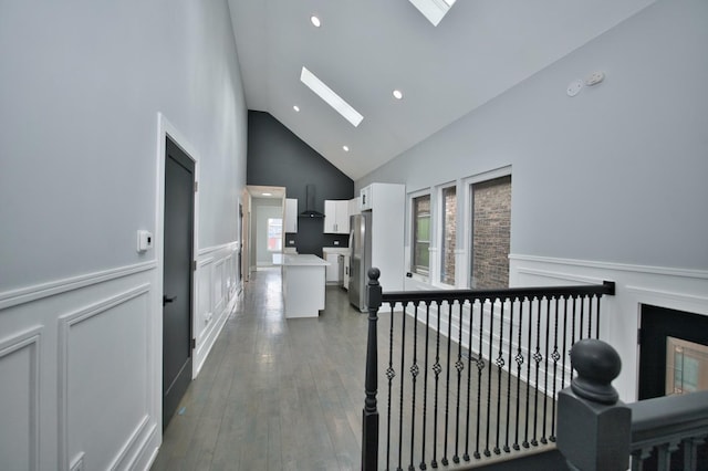 corridor with a wainscoted wall, dark wood finished floors, a skylight, a decorative wall, and high vaulted ceiling
