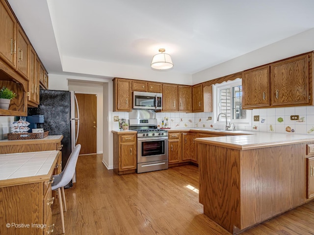 kitchen with light wood-style floors, brown cabinetry, stainless steel appliances, a peninsula, and backsplash